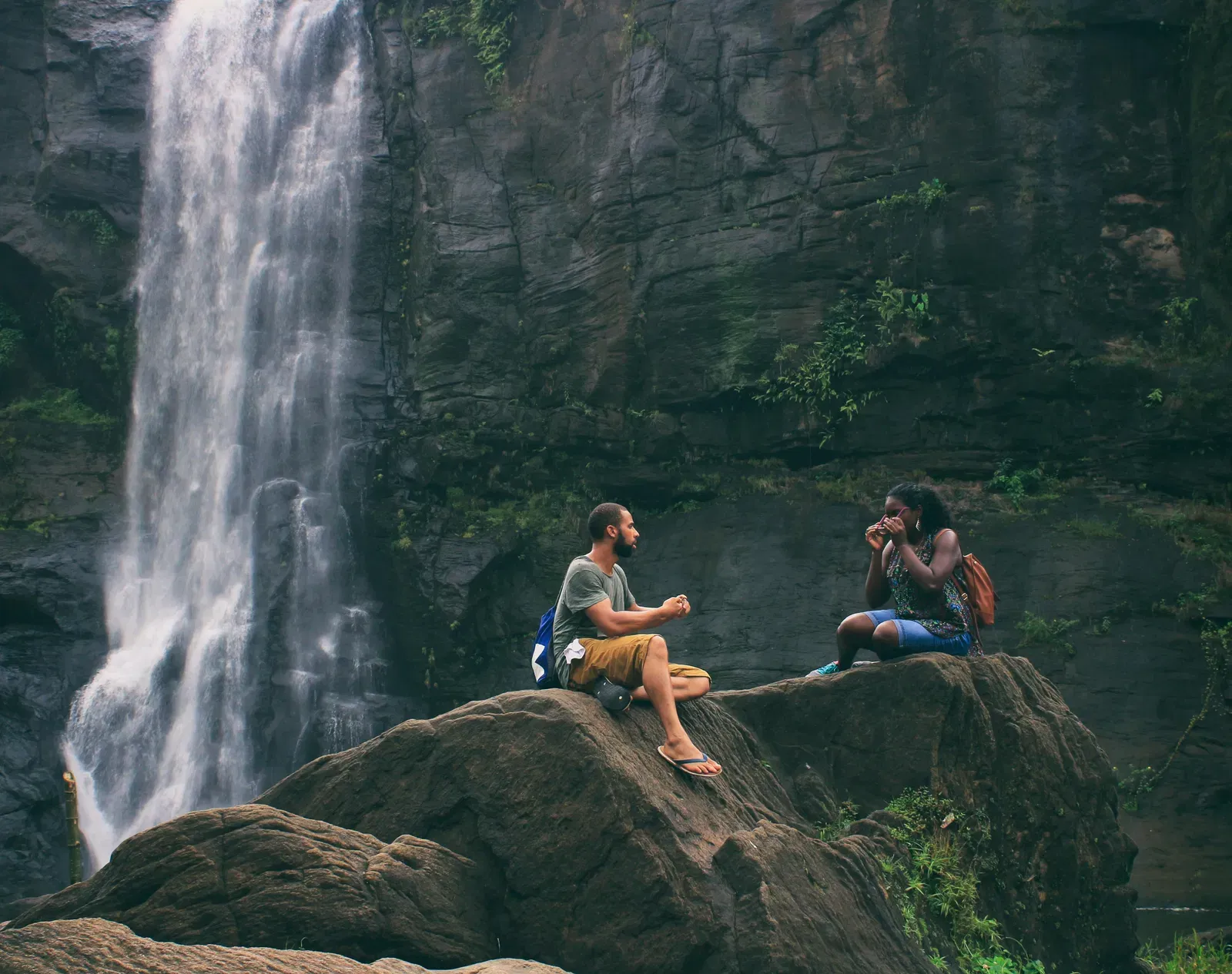 Two people sitting on a rock cliff in front of a waterfall.
